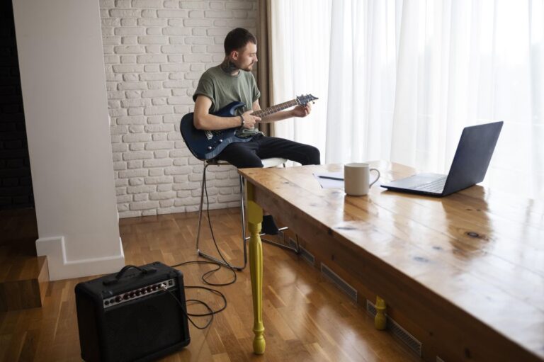 Man practicing electric guitar at home beside a window, with an amplifier on the wooden floor and a laptop on the table